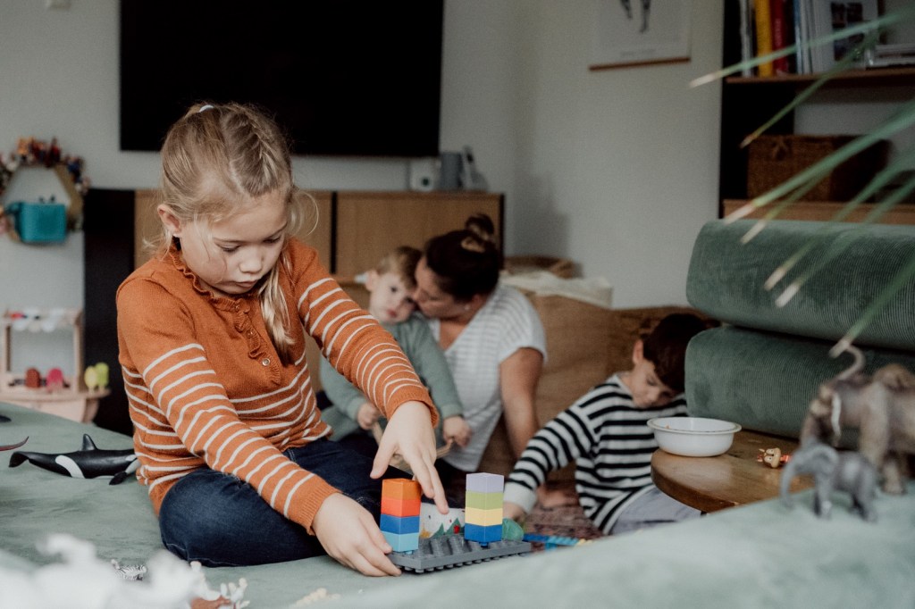Kleines Kind spielt mit Lego während hinter ihr Bruder mit Schleichtieren spielt und die Mama ihr jüngstes Kind tröstet