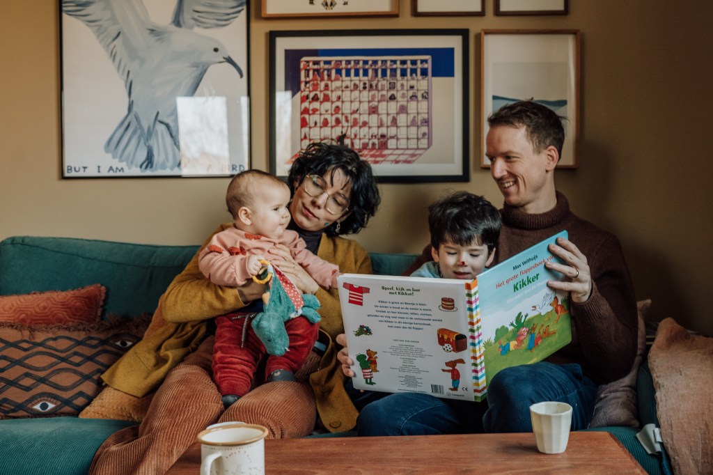 Mama hält Baby auf dem Arm während Papa mit Kleinkind ein Buch liest