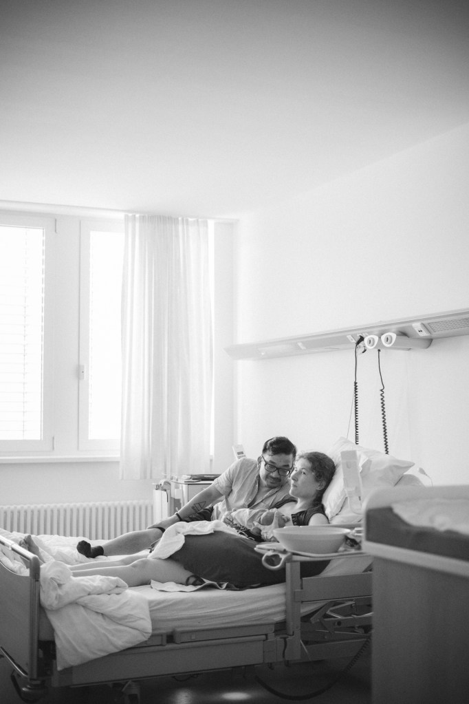 Authentische Wochenbettfotografie in Freiburg: Eltern kuscheln ihr Neugeborenes im Krankenhausbett im Projekt Mutter werden von Thuy Hang Willmann Fotografie.